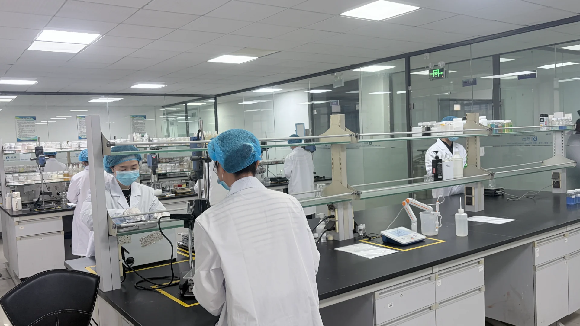 Scientists in lab coats and hair covers working at lab stations in a modern research laboratory with glass partitions and equipment.
